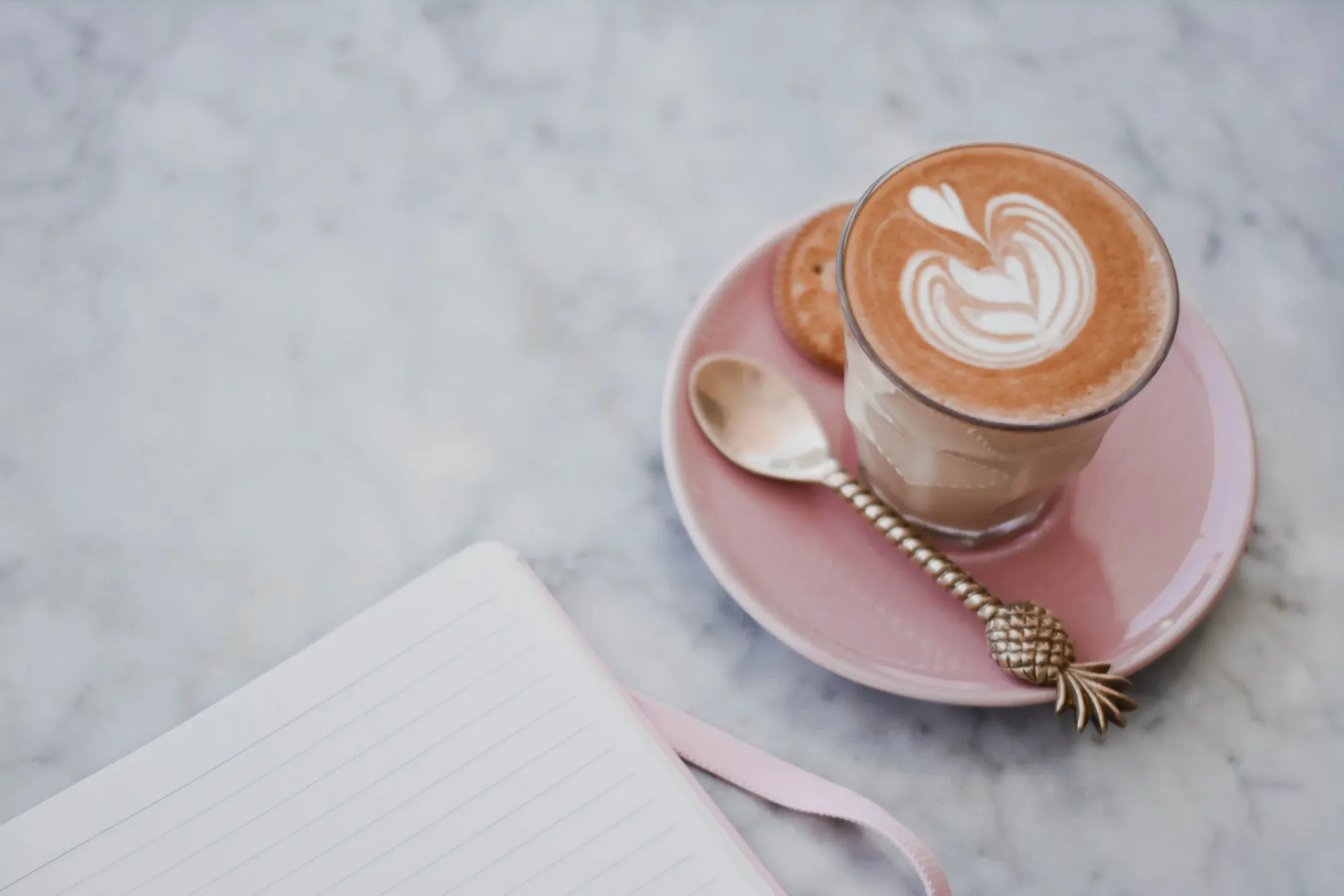 cappucino inside a glass mug on pink saucer next to a notebook on a marble table
