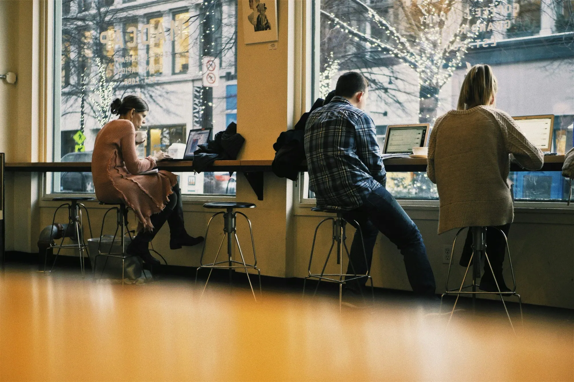 3 people sitting along a coffee shop window working on laptops
